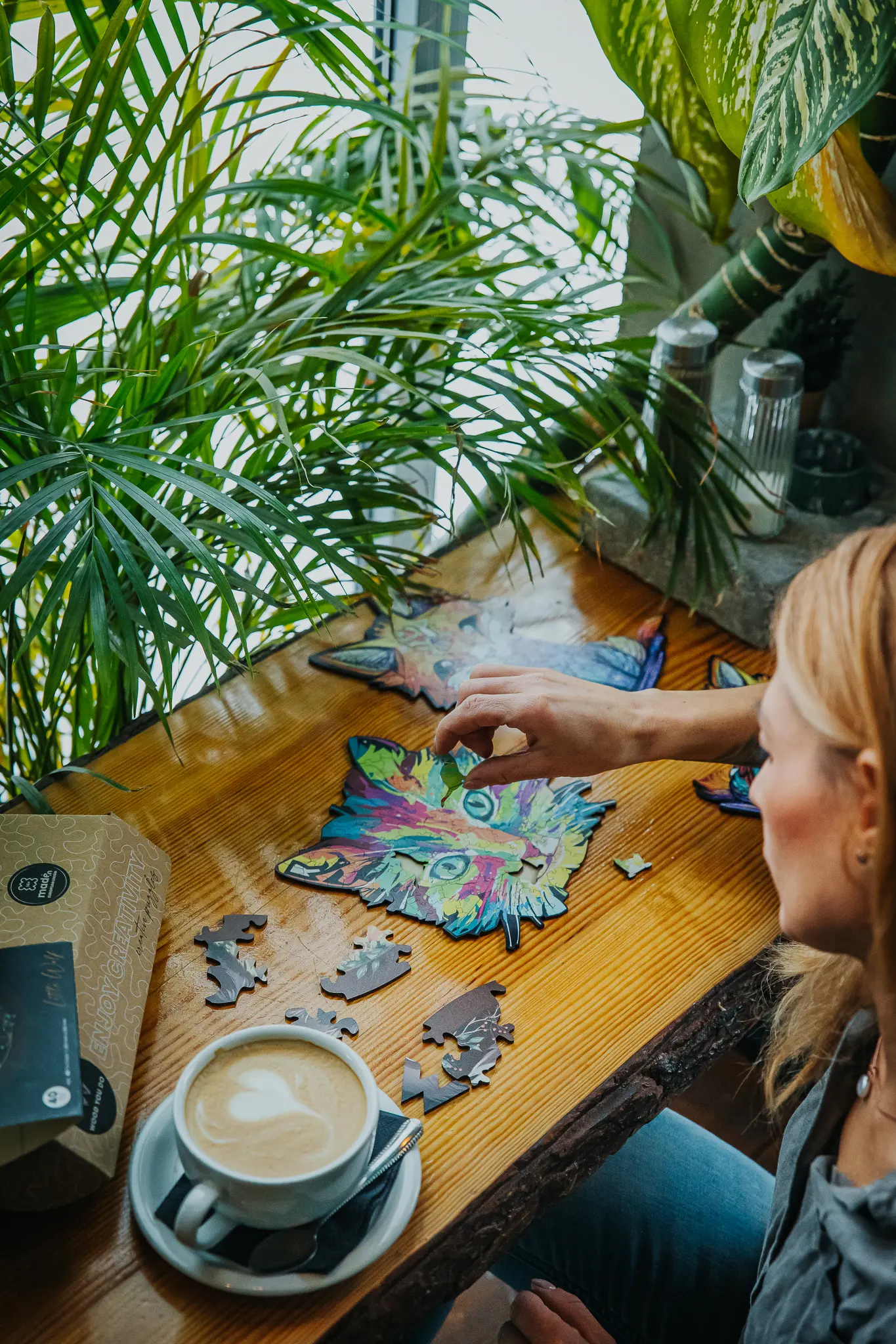 Kobieta siedzi na chokerze przy restauracyjnym blacie i układa drewniane puzzle w kształcie rysia. Obok niej stoi filiżanka z kawą.