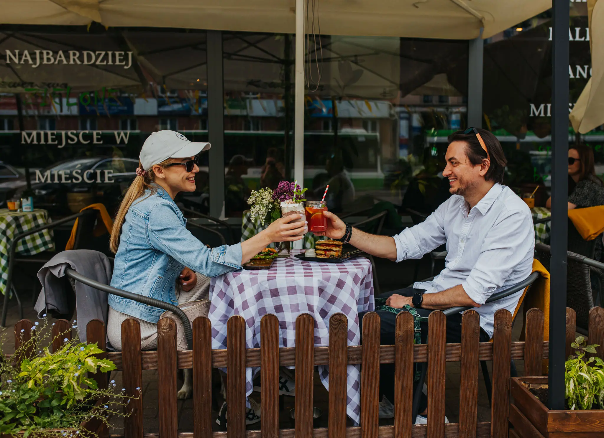 W ogródku restauracyjnym siedzi para, piją koktajle i wznoszą toast.