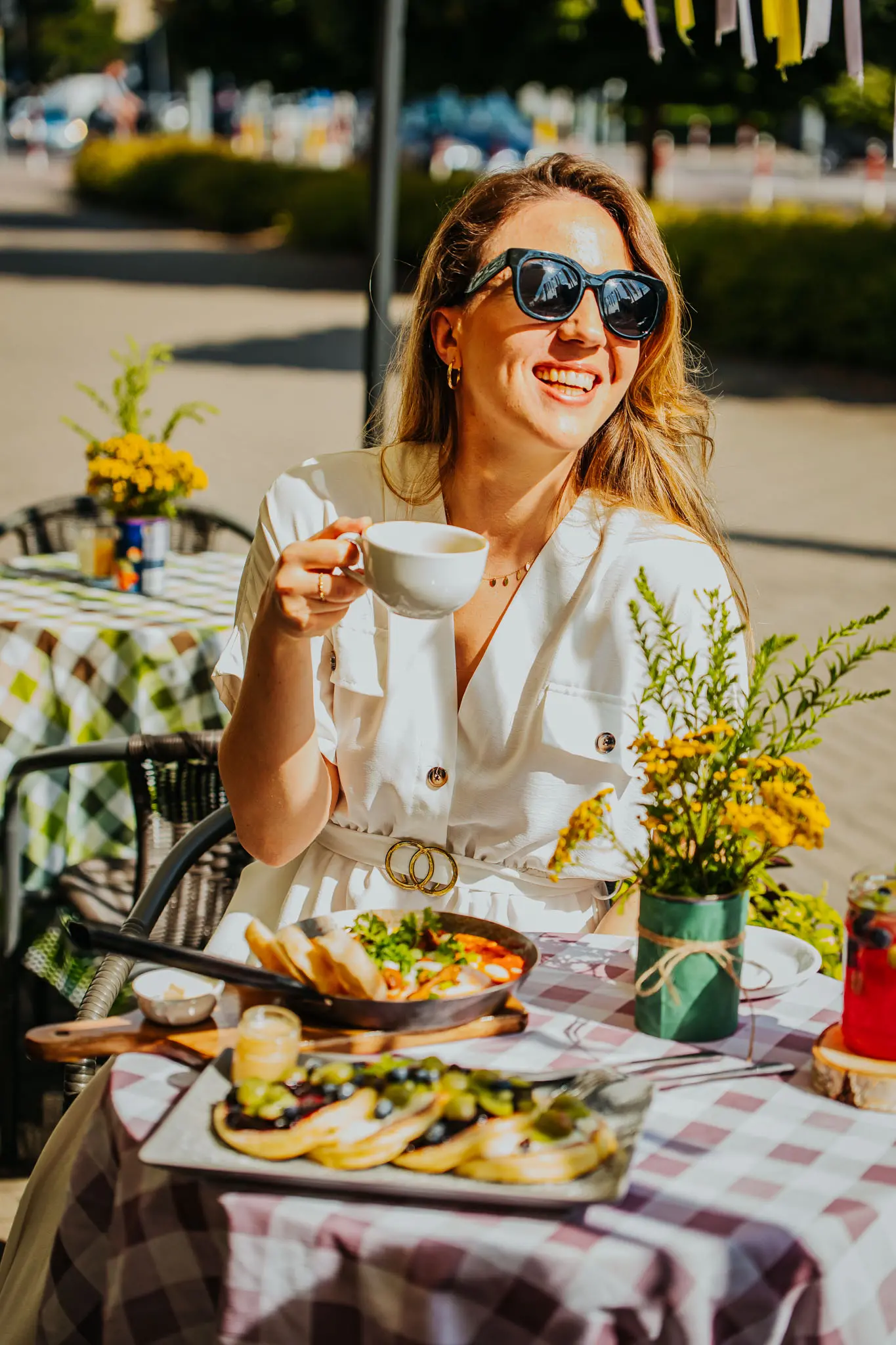 Kobieta o blond włosach, ubrana na biało w okularach przeciwsłonecznych spożywa posiłek w ogródku restauracyjnym, uśmiecha się a w dłoni trzyma filiżankę kawy.