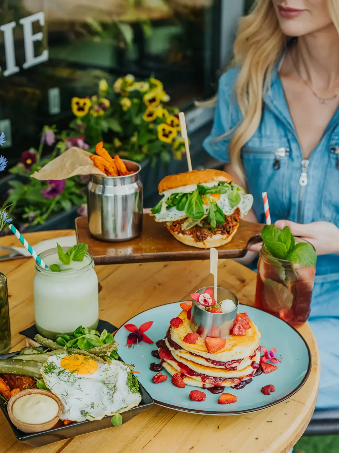 Kobieta siedzi przy stole w ogródku restauracyjnym, który jest pełen dań: naleśniki z truskawkami, letnie koktajle, burgery, zestaw śniadaniowy i frytki